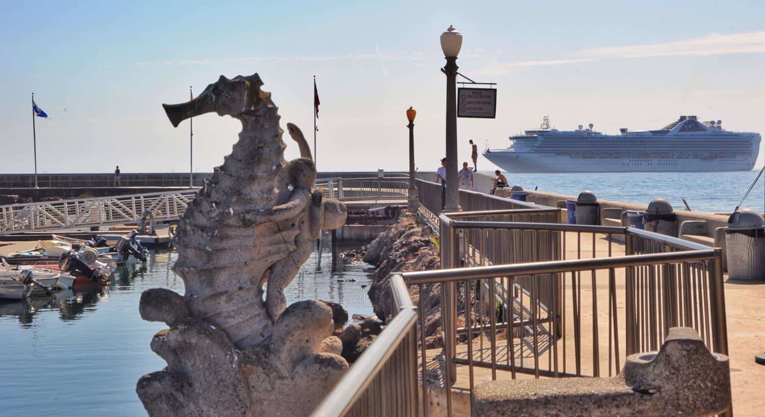 Seahorse sculpture in the harbor with docked boats and a cruise ship in the background