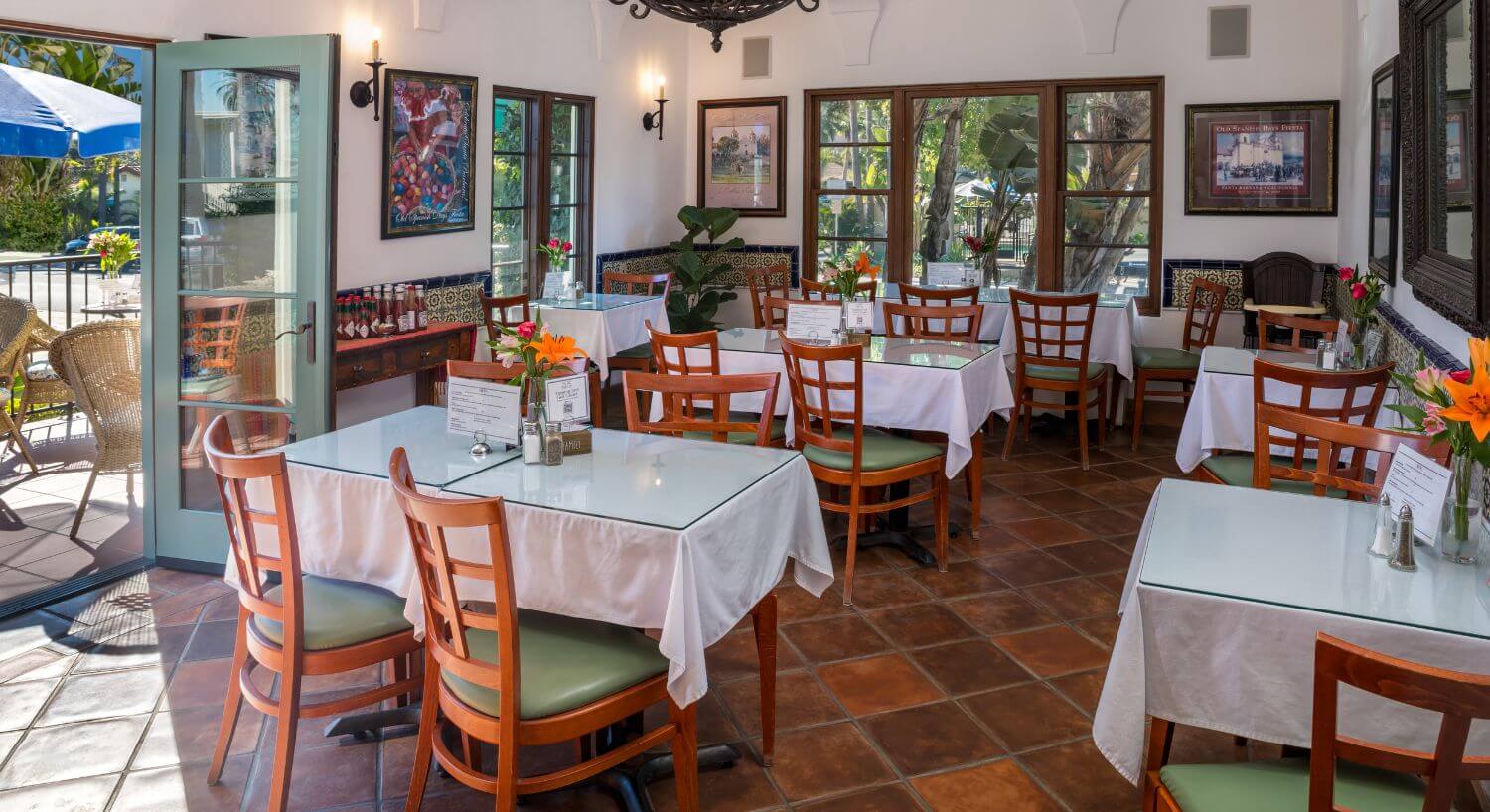 multiple rectangular wood tables with glass tops over white linen tablecloths, green cushioned wood chairs, patio door open