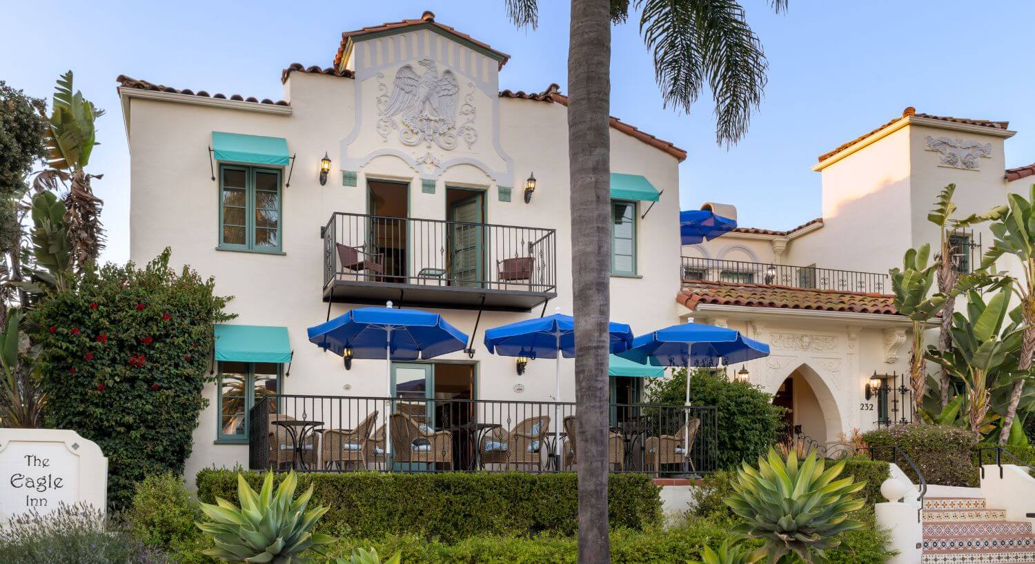 two cream two-story buildings separated by 2nd floor balcony, the eagle inn sign, patio with wicker furniture and blue umbrellas