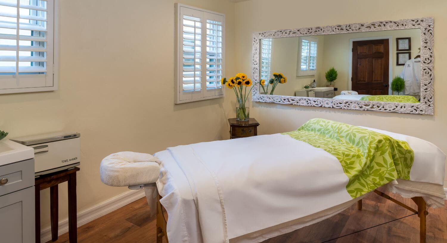 cream painted room with massage table, windows with shutters, sunflowers in vase in corner, rectangular mirror on wall