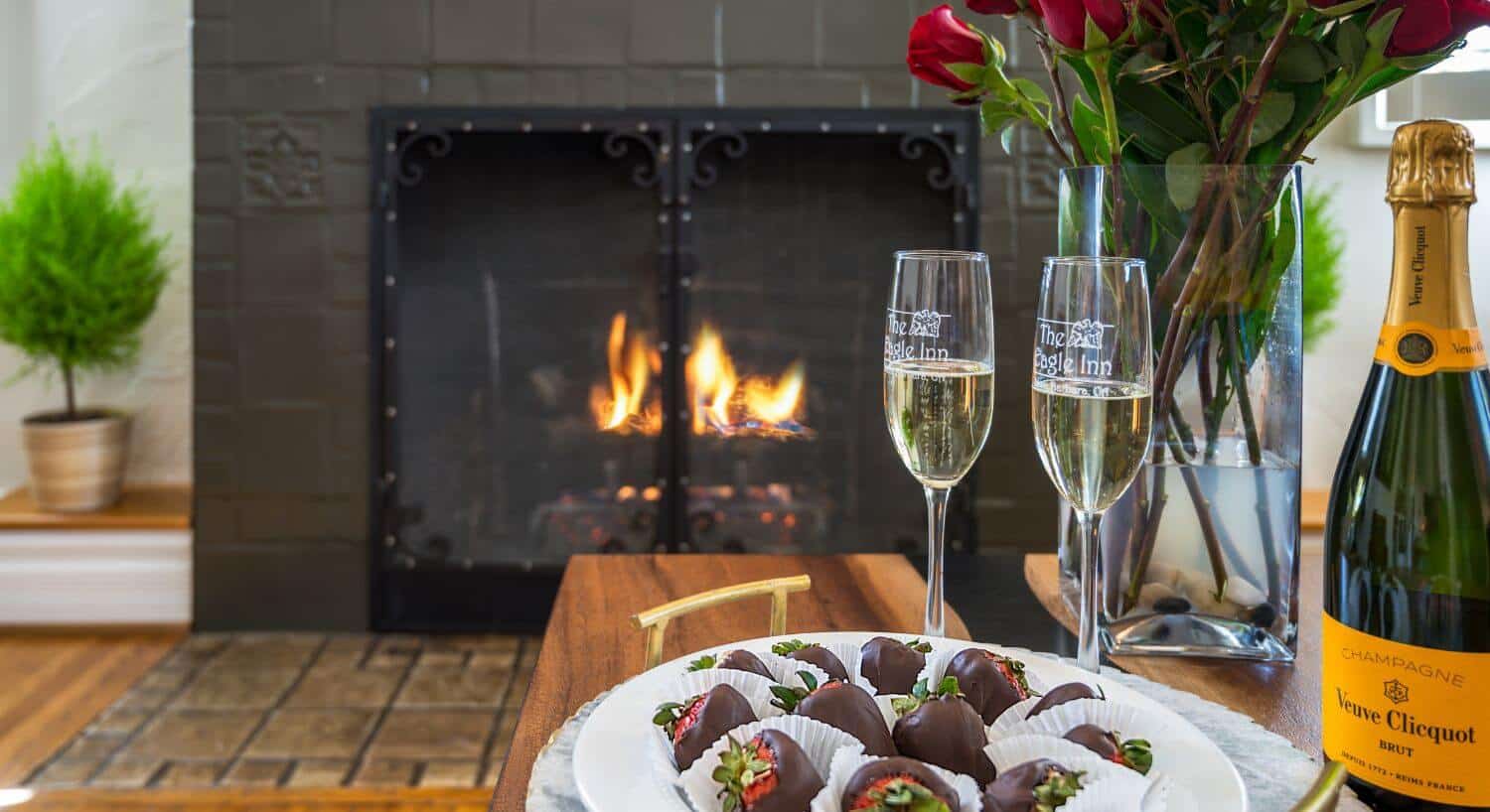 two champagne glasses with the eagle inn logo, tray of chocolate covered strawberries, red roses in vase, fire burning in fireplace behind