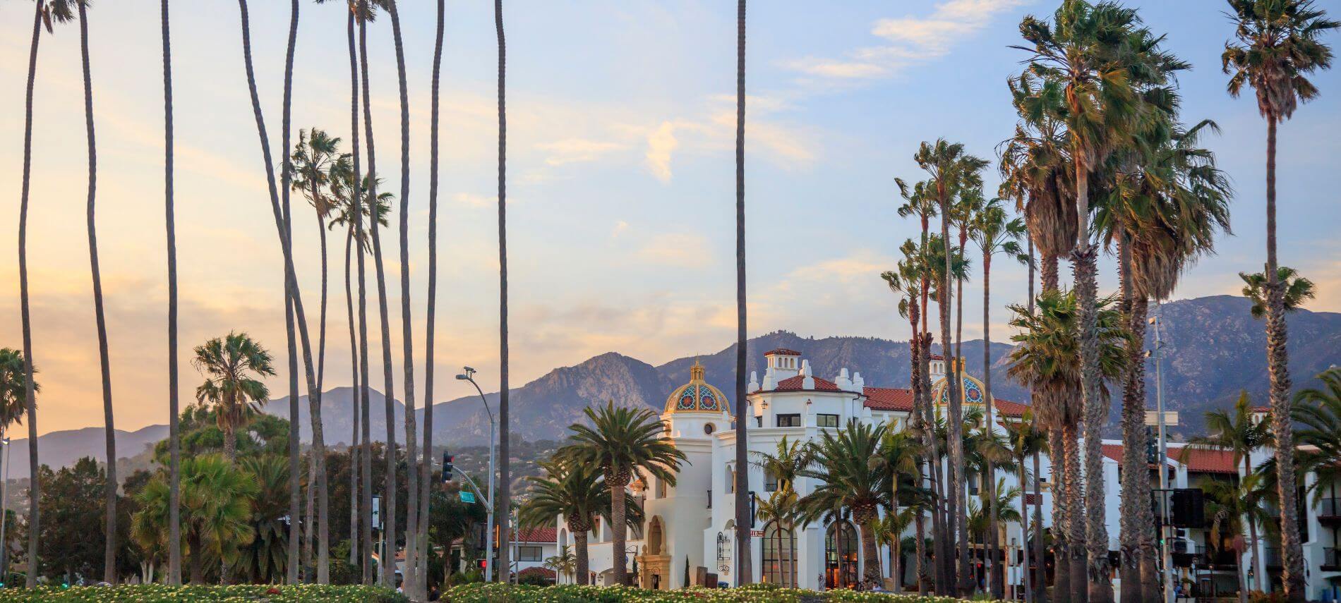 Palm lined street with Spanish style buildings and mountains in Santa Barbara at sunset.