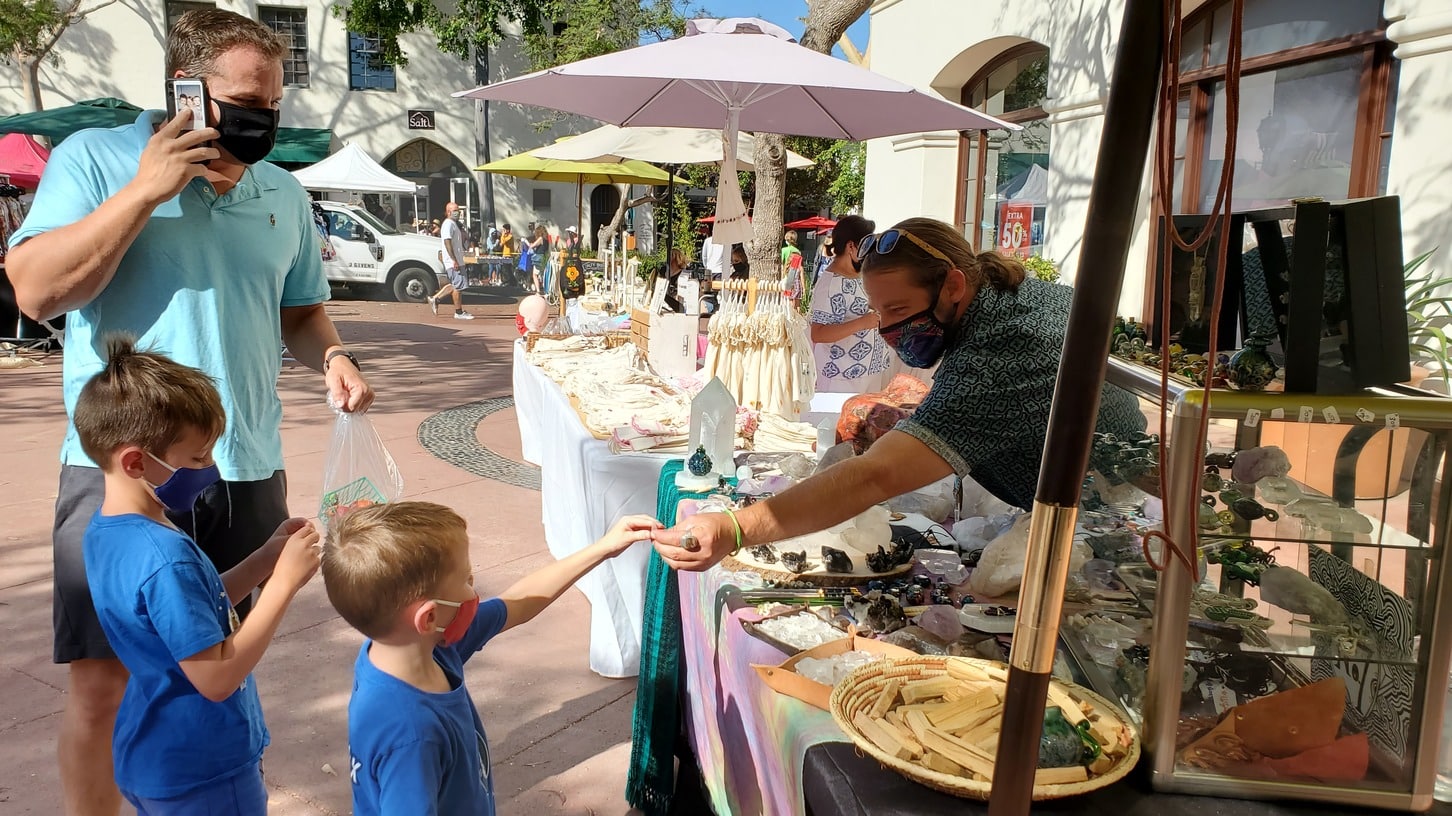 A craftsman is leaning over a table of his products, handing a sample to a young boy in an outdoor marketplace.