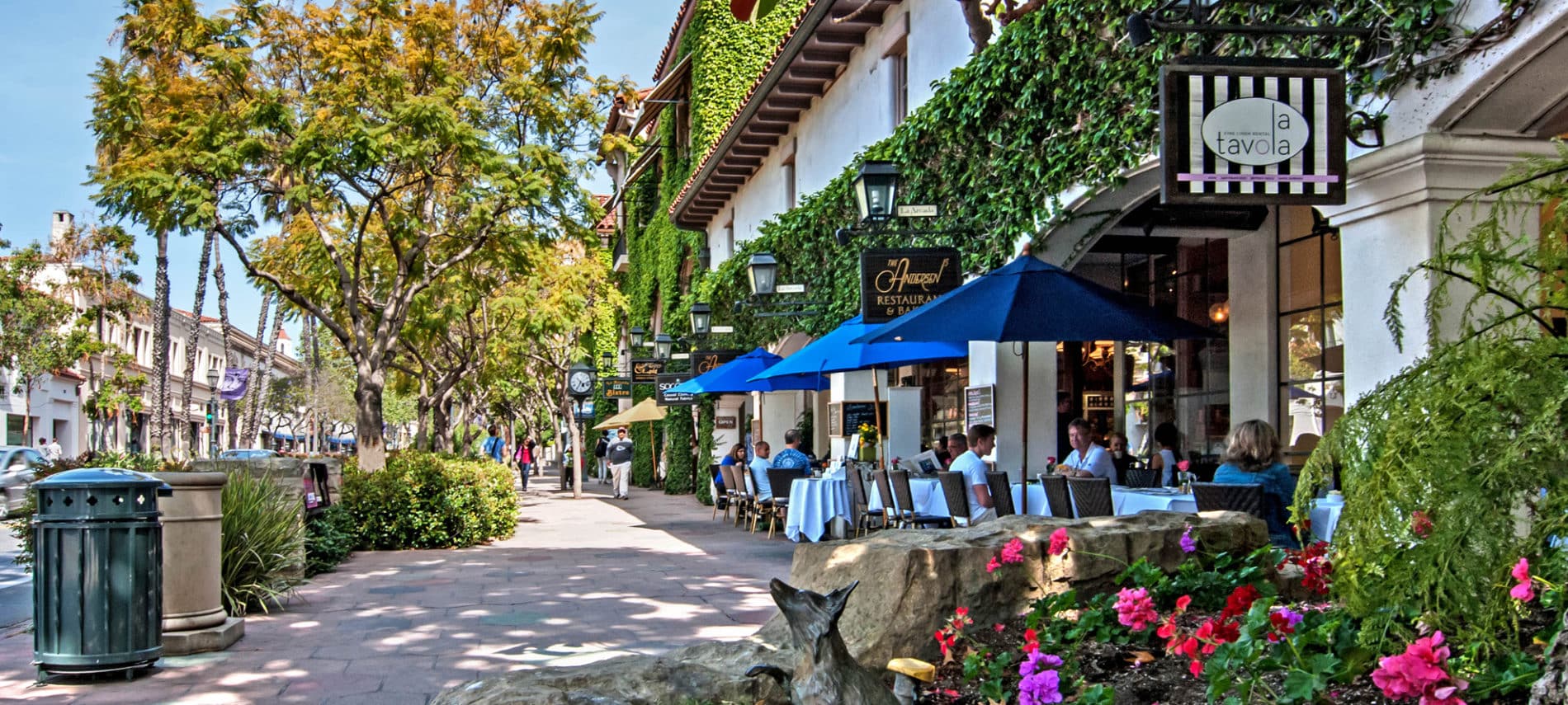 A sidewalk with bright flowers in the foreground, a Spanish-style restaurant with red roof and diners eating under blue table umbrellas.