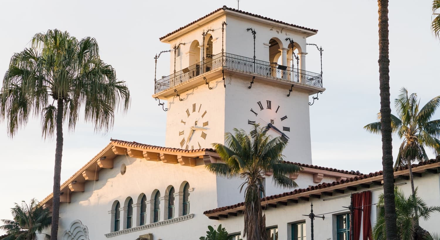 Spanish style ivory stucco building with red tile roof, large clock and tops of palm trees
