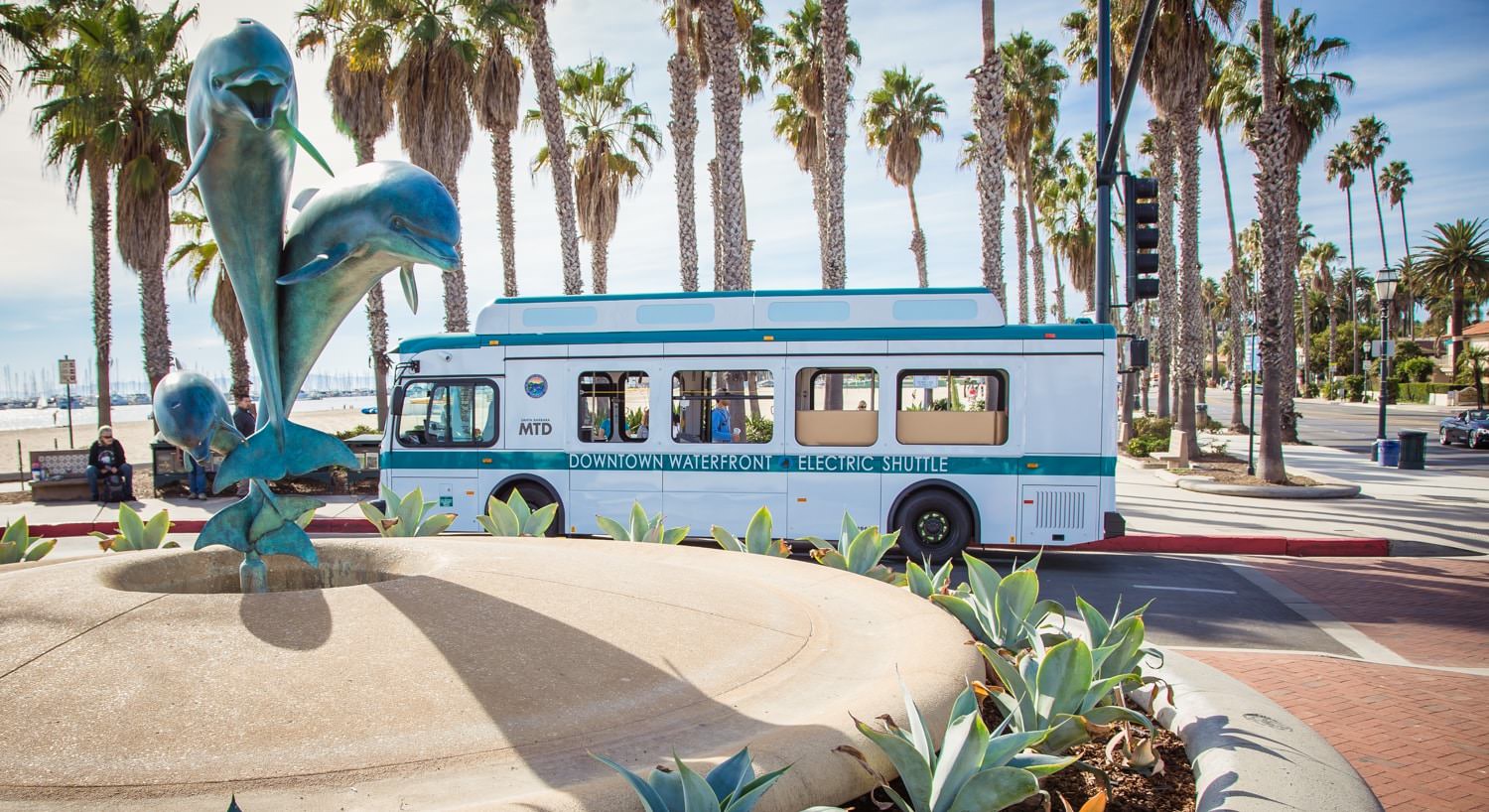 White and turquoise shuttle bus riving around Santa Barbara amidst palm trees and harbor views