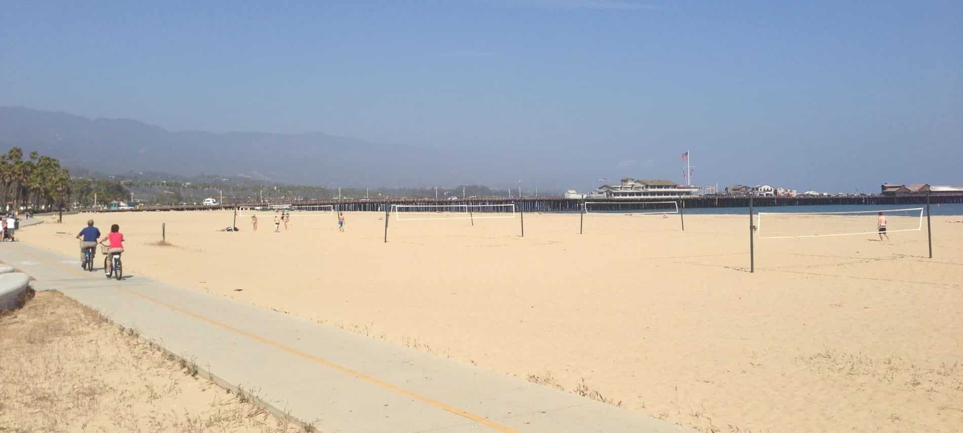 People riding bikes along a sandy beach with volleyball nets surrounded by mountains