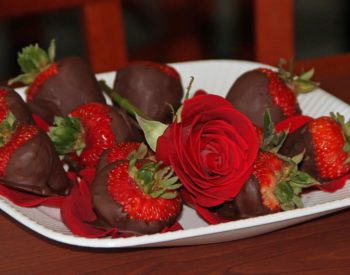 White tray topped with red strawberries with green stems dipped in chocolate on a bed of red rose petals and one red rose