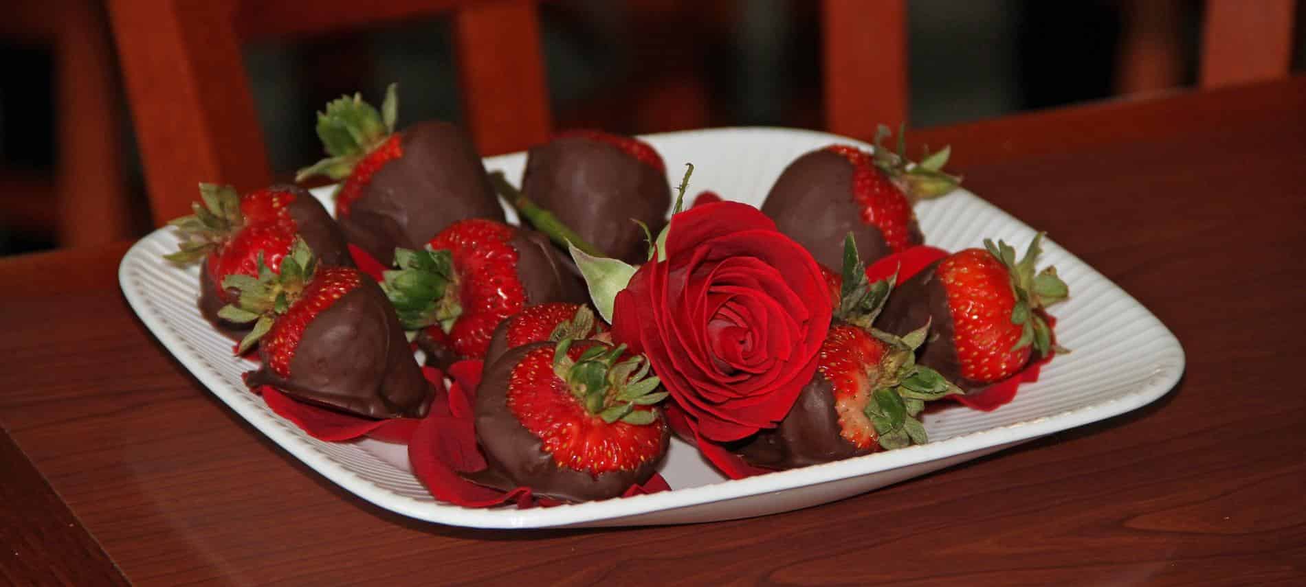 White tray topped with red strawberries with green stems dipped in chocolate on a bed of red rose petals and one red rose