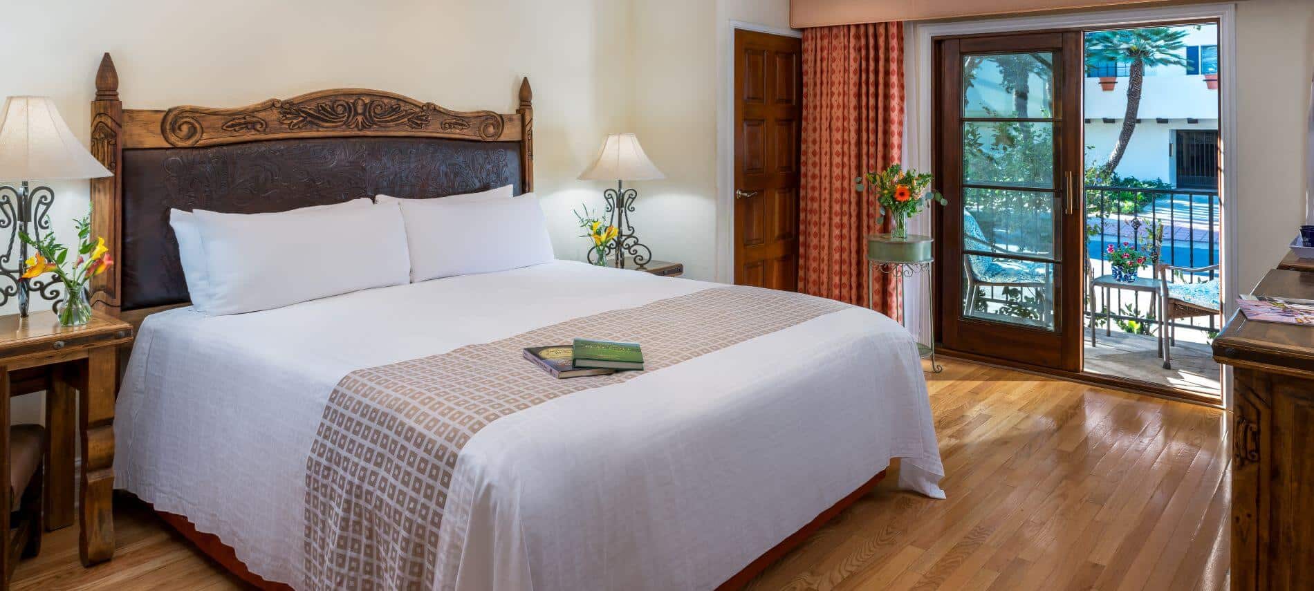1st floor cream room with king bed with ornate headboard, white bedspread, stands with lamps, wood floor, private patio