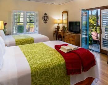 tan room with two queen beds with white bedspreads, dresser with tv on top, desk in corner with lamp, door open to private patio