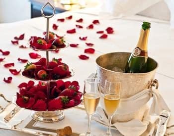 Table topped with red rose petals, three-tiered plates with strawberries, two flutes, and bucket of chilled champagne