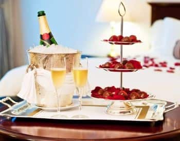 Silver tray on a round table topped with chilled champagne, two full champagne flutes and three tiered tray with goodies