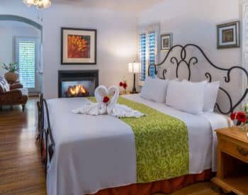 well lit cream room with king wrought iron bed, brown loveseat close to fireplace, stands with lamps, towels formed like swans