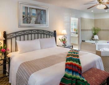 well lit carpeted white room with king bed, colorful blanket across end of bed, tub in corner, ceiling fan, stands with lamps