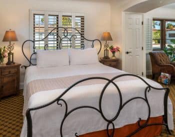 wrought iron queen bed in white carpeted room, shuttered windows, stands on side, separate sitting area