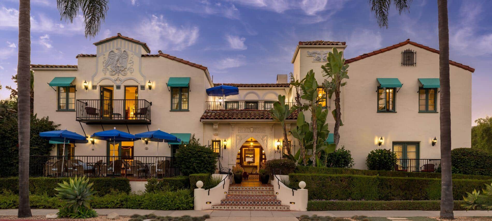 front view of two cream buildings separated by stairs into lobby, balcony and patio with tables and blue umbrellas, steps leading into building