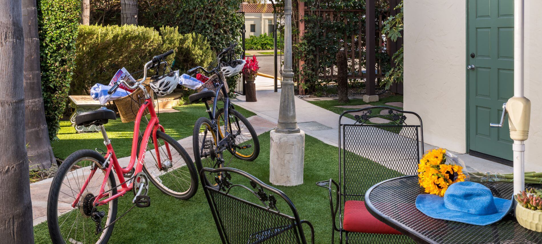 red and blue bikes sitting on grass by sidewalk and lamp post, metal table and red cushioned chairs under blue umbrella, flowers on table