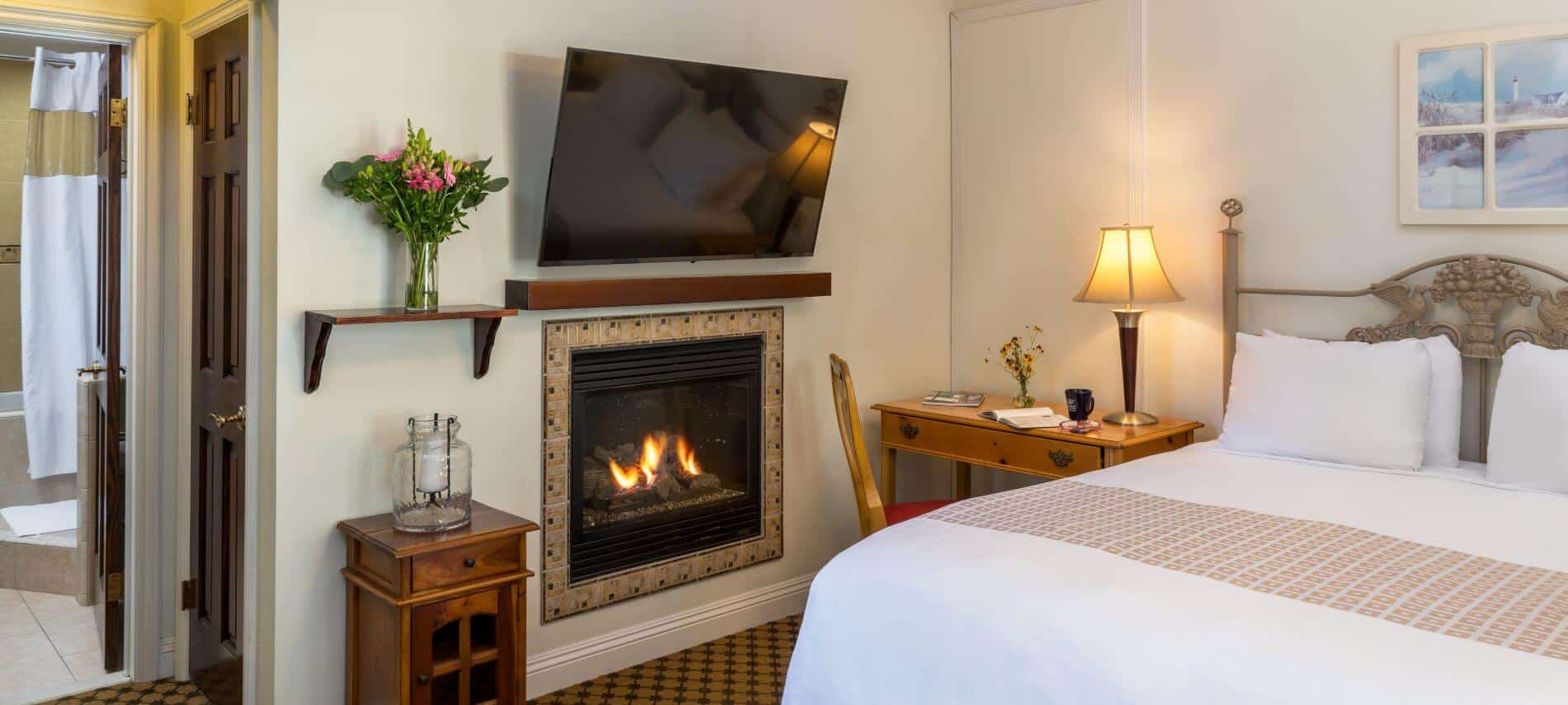 cream room with queen bed, desk with lamp, flat screen tv on wall over fireplace.