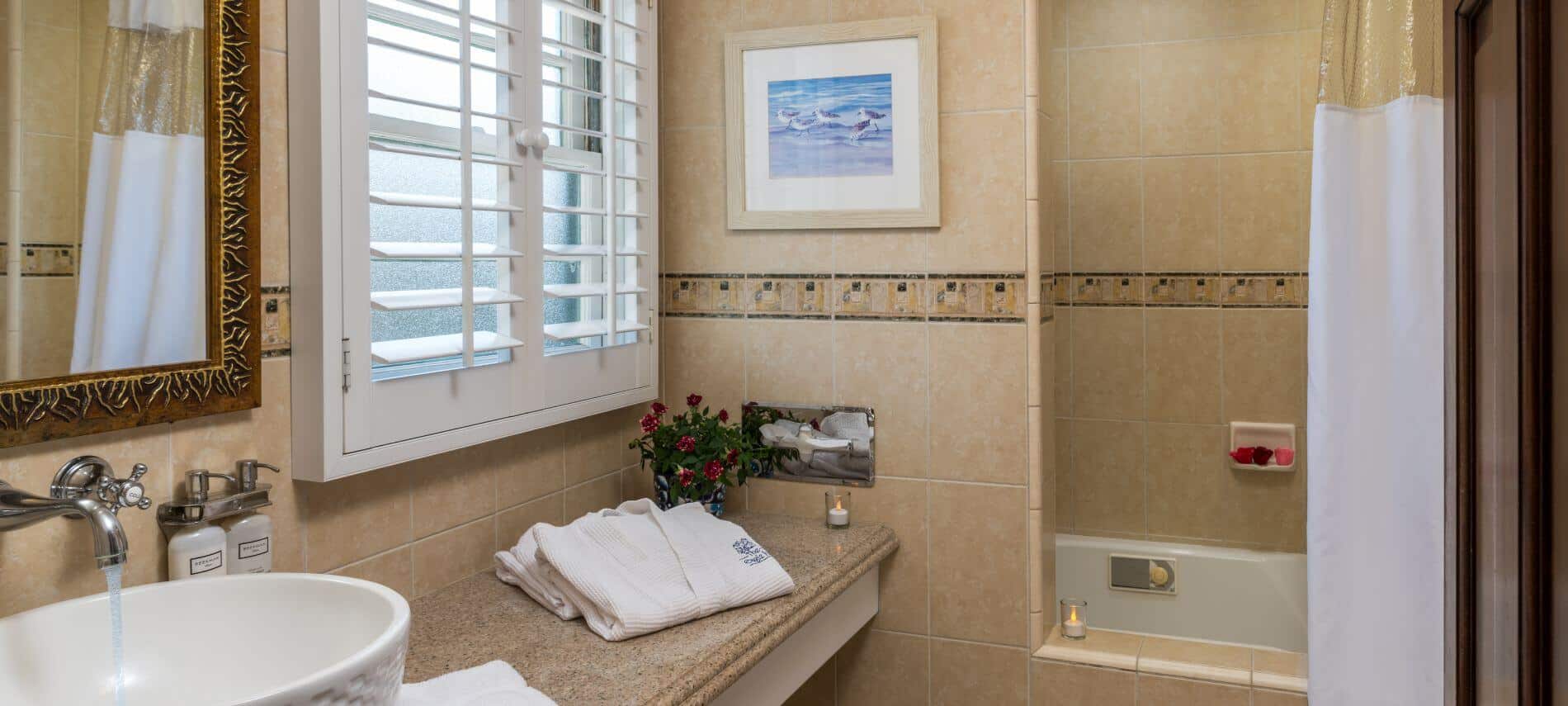 well lit tiled bathroom with long counter with sink on top and gold mirror, window with shutters, shower