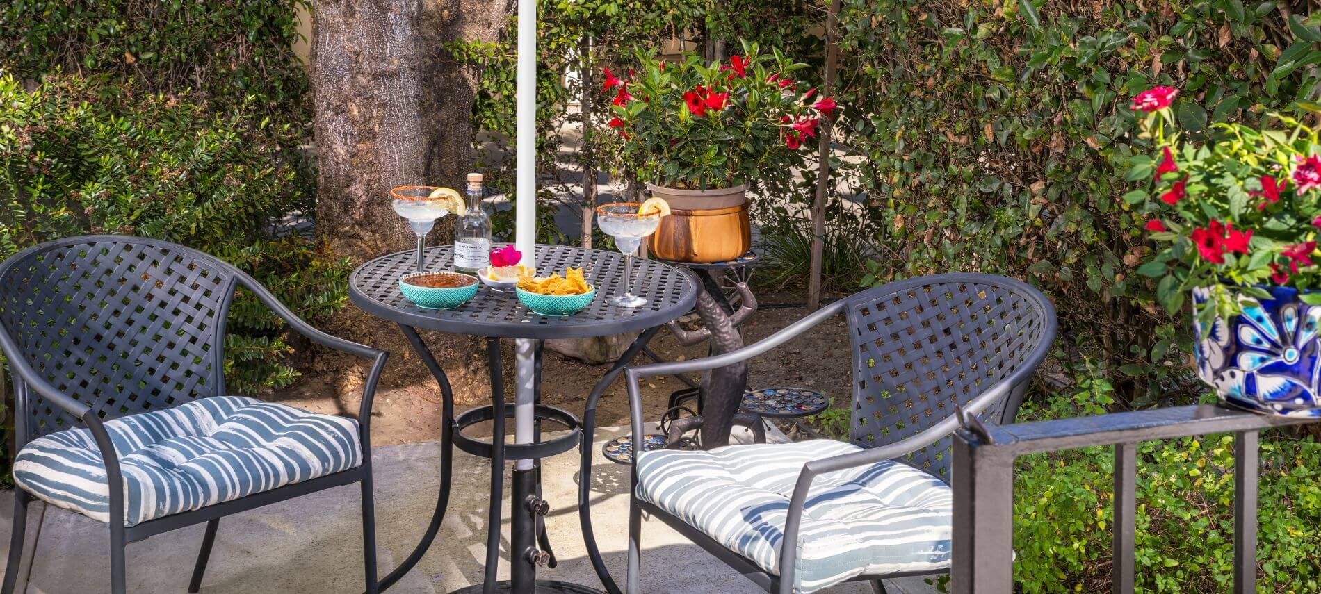 outdoor round metal table and cushioned chairs under blue umbrella in garden with flowers in bloom
