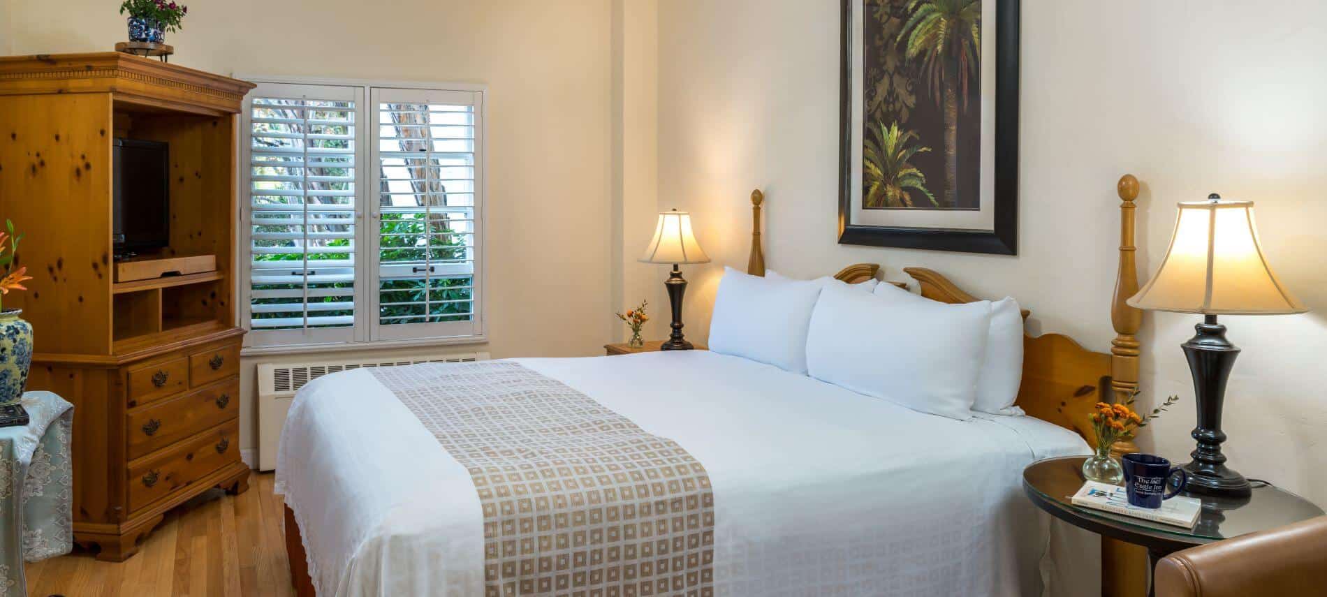 small cream room with king bed, wood floors, stands with lamps, flat tv in corner hutch