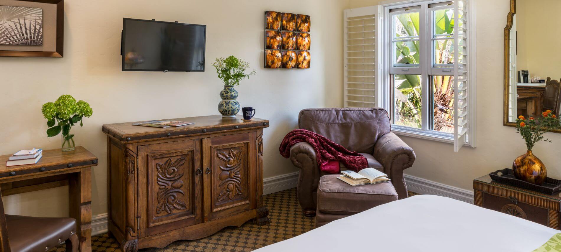 tan room with view across bed toward window with brown chair in corner, flat tv on wall, oak cabinet