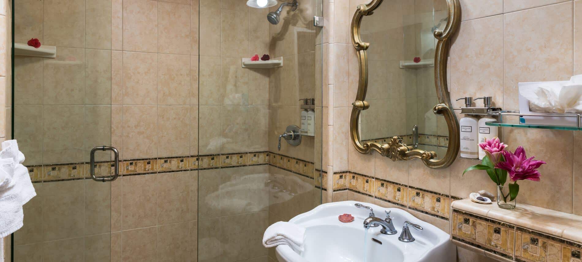 tiled bathroom with white sink with running water under gold trimmed mirror, white cloth on edge, glass walk-in shower
