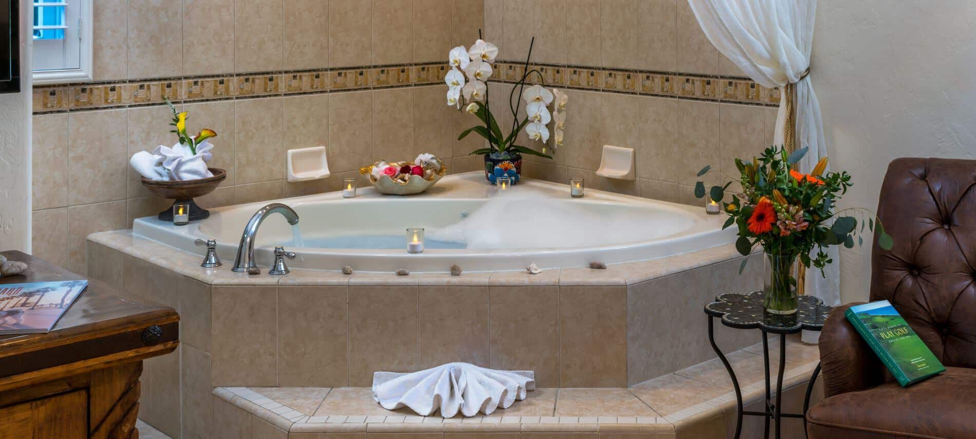 corner tub with bubbles in tiled walls, brown chair by tub next to stand holding flowers