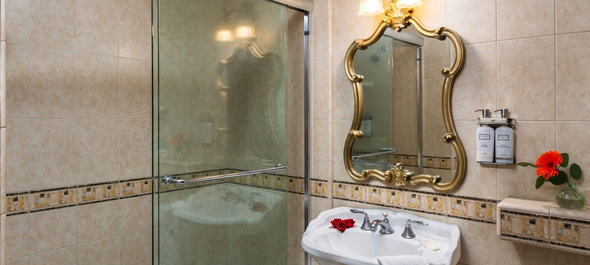 tiled bathroom with walk in shower, sink with gold trimmed mirror hanging above, dispensers with lotion and soap on wall