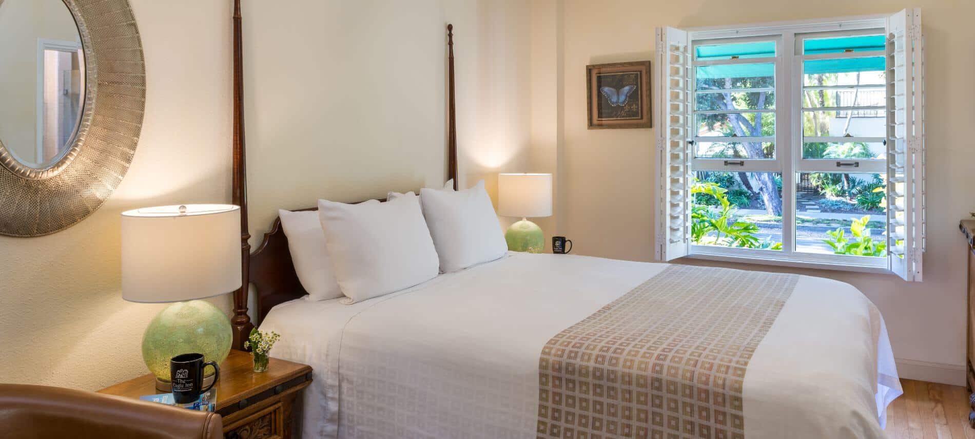 cream 1st level room with queen two-poster bed by street with open windows, stands with lamps, mirror on wall, wood floors