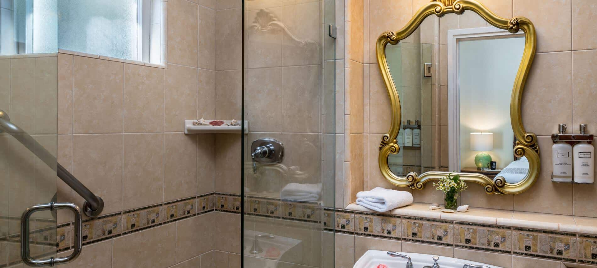 tiled bathroom with sink with gold mirror hanging over, walk in glass enclosed shower with grab bar