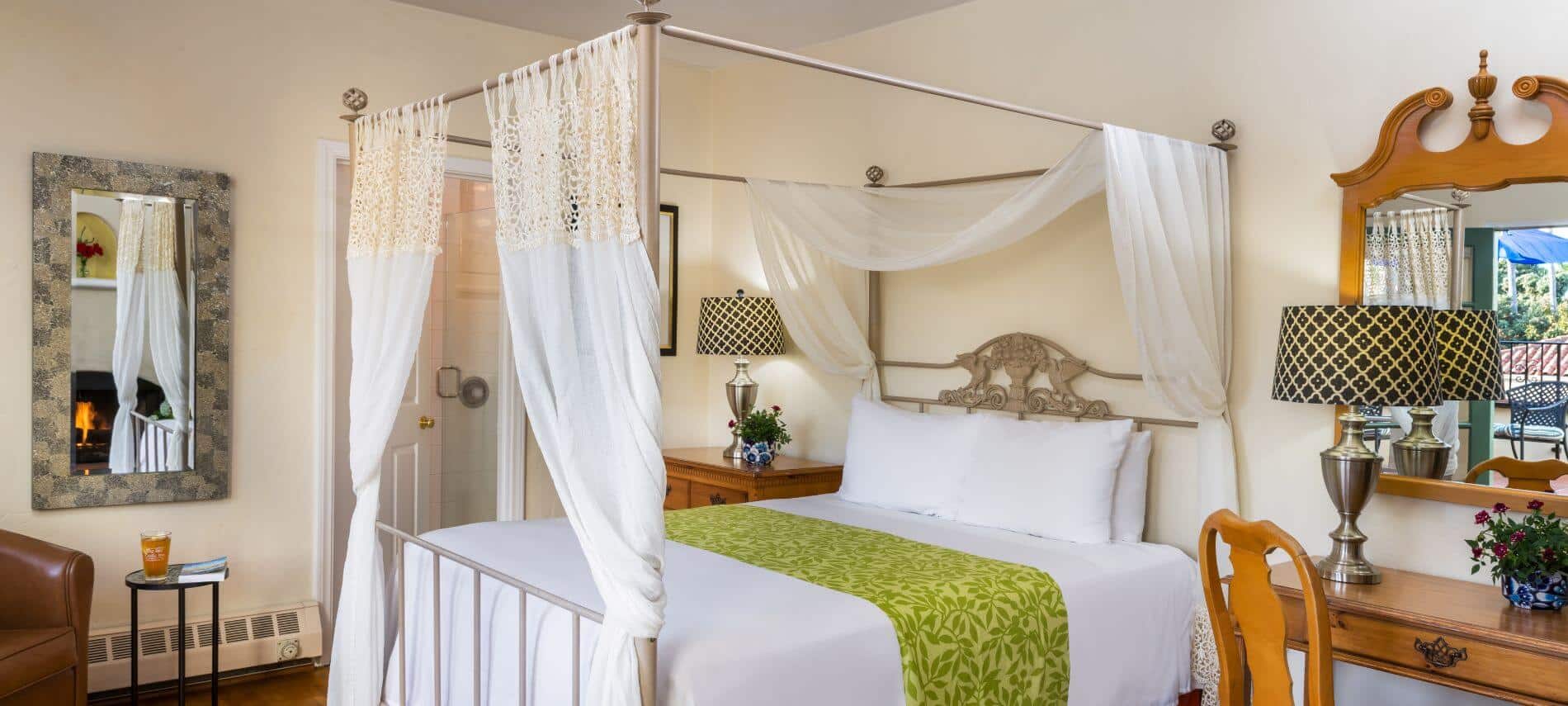 wood floored cream room with queen canopy bed with white bedspread, desk with lamp and red cushioned chair with mirror above