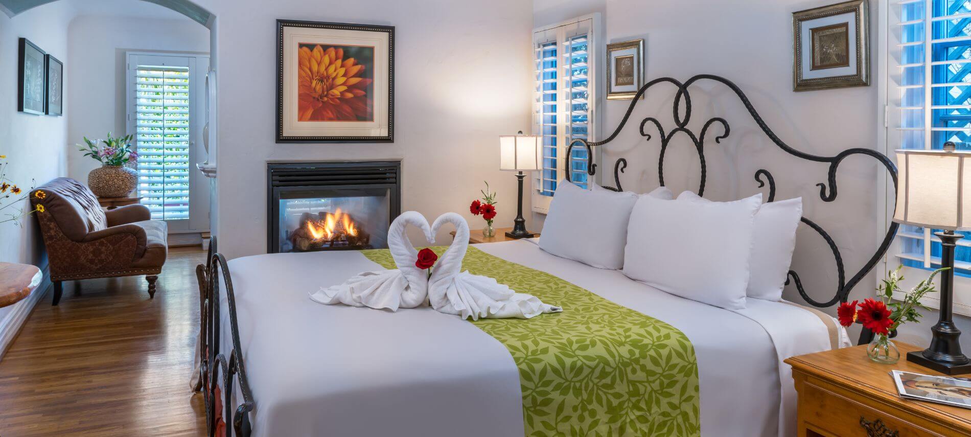 well lit cream room with king wrought iron bed, brown loveseat close to fireplace, stands with lamps, towels formed like swans