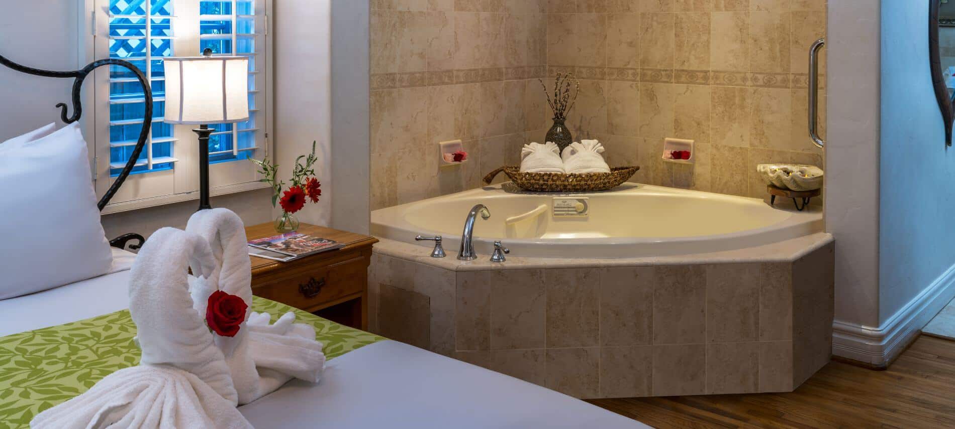 bathtub in tiled corner of room, bed with towels in shape of swans, stand with lamp by window