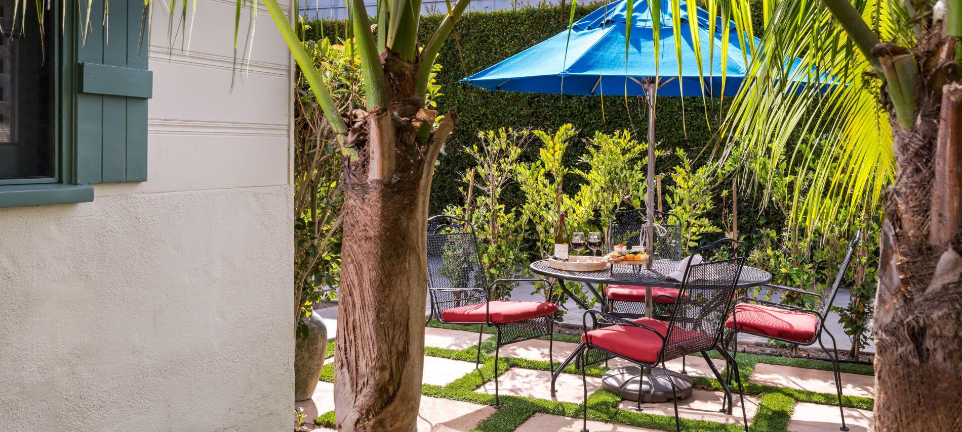 black round metal table with red cushioned chairs under blue umbrella in garden by tan house