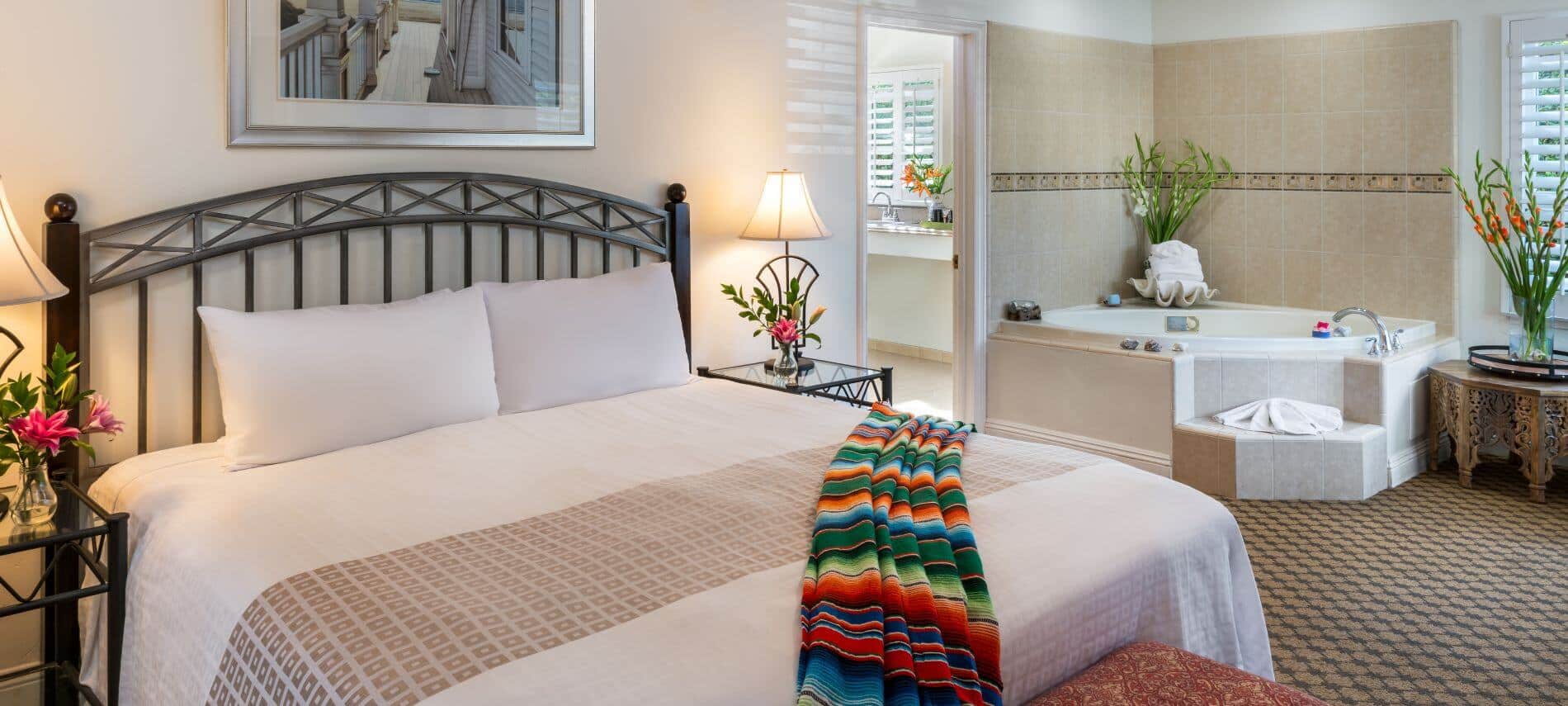 tan carpeted room with king bed, stands with lamps, tiled corner of room with tub and steps to enter