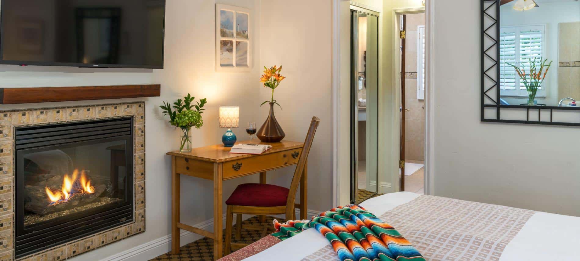 tan room with fireplace, flat tv, desk, view across bed with colorful blanket on end with connecting bathroom