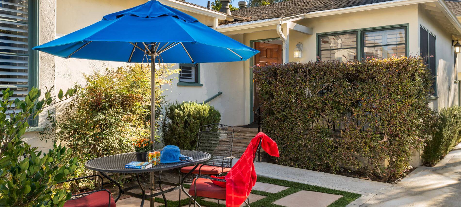 back of tan single story house, round metal table and red cushioned chairs under blue umbrella in garden setting