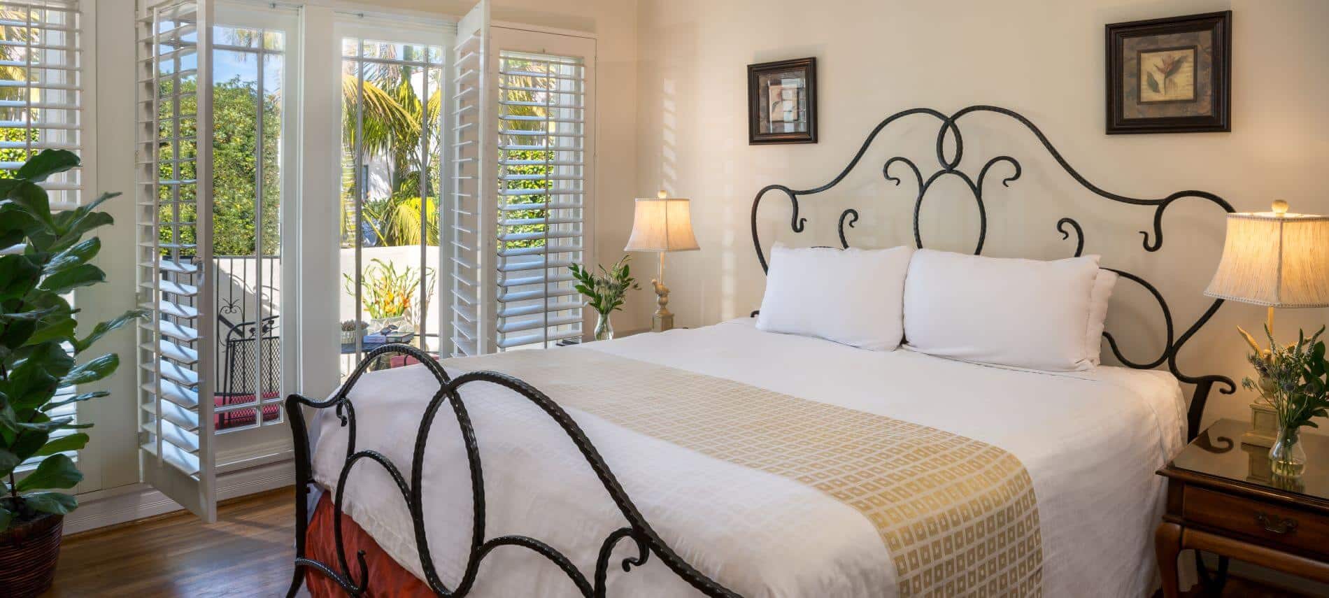 side view of wrought iron king bed in tan room with wood floor, stands with lamps, private patio, artwork on wall
