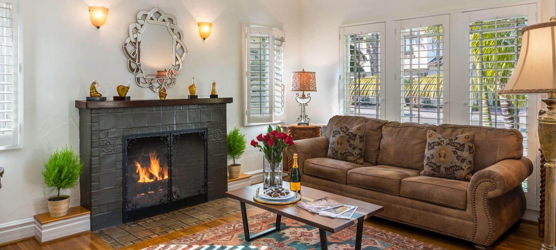 white living room with brown couch, coffee table holding refreshments, fireplace under silver framed mirror, shuttered windows, standing lamp