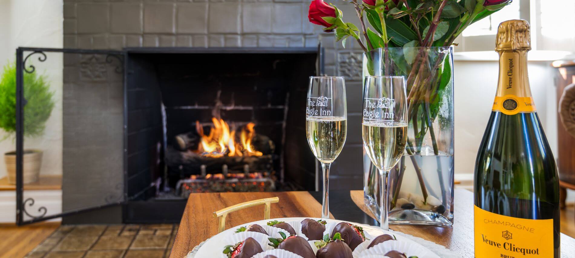 champagne glasses and bottle sitting by plate holding chocolate covered strawberries, vase with red roses, fireplace burning