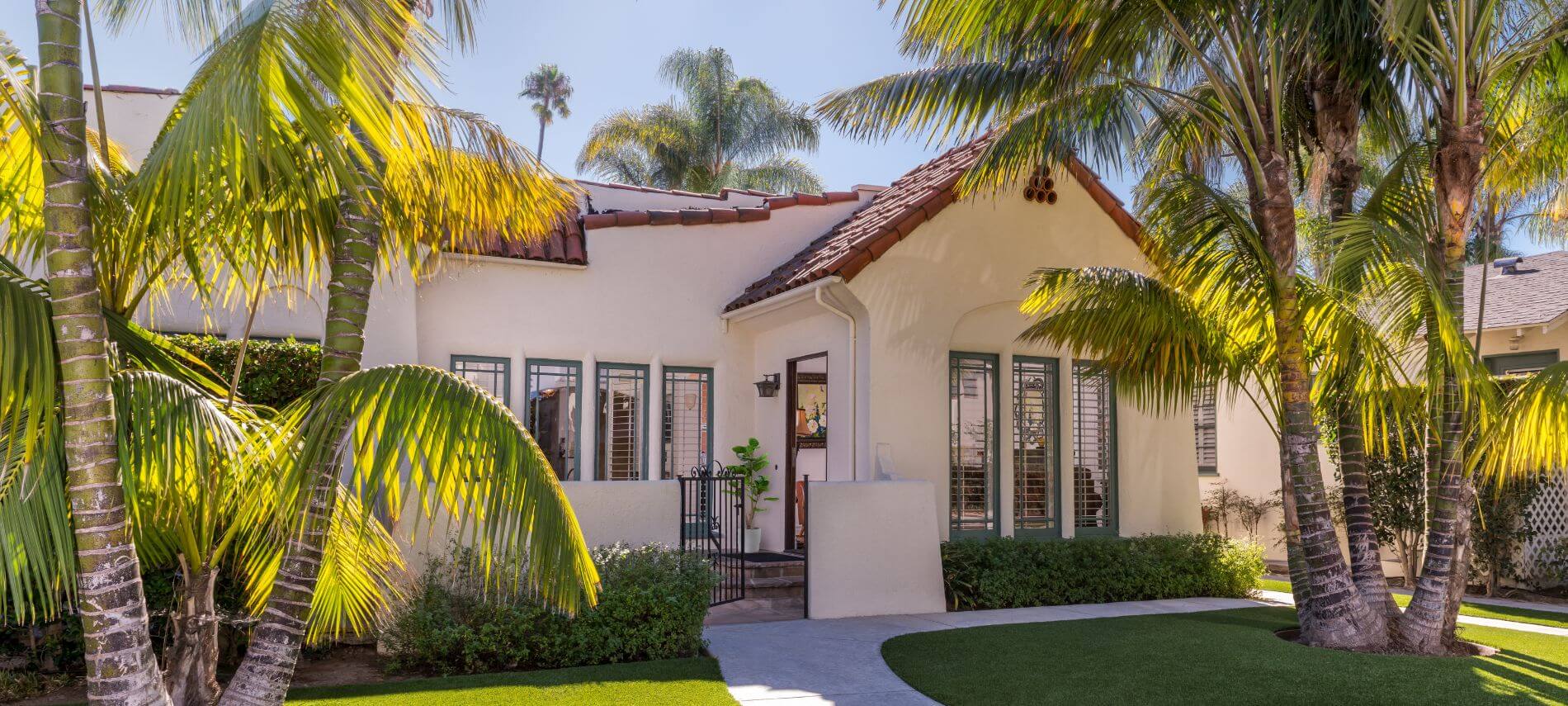 small tan single story house with clay roof surrounded by palm trees, shuttered windows