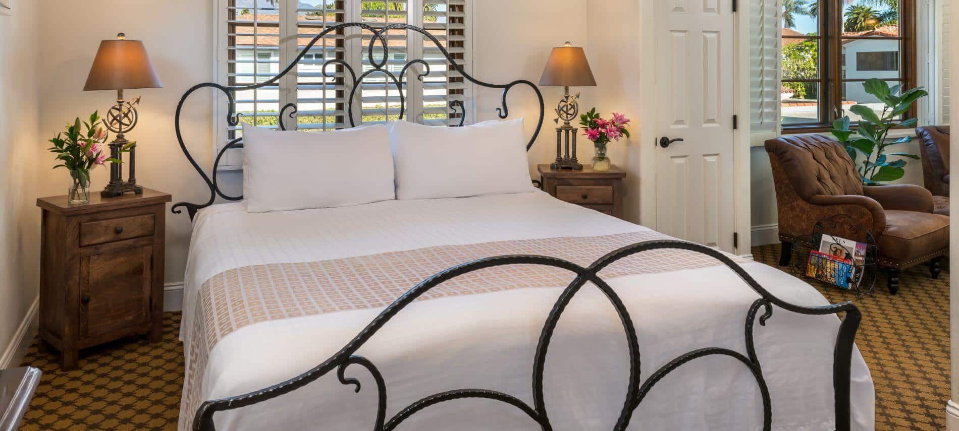 tan carpeted room with wrought iron queen bed, stands on both sides with lamps, sitting room with brown chairs