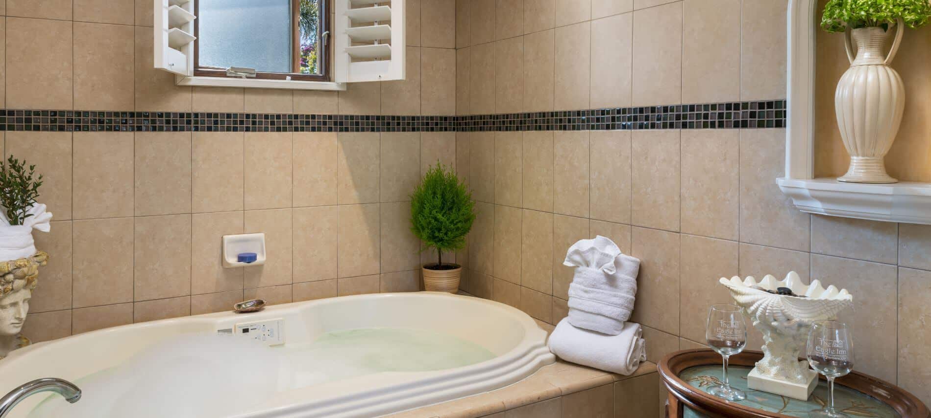 tan tiled bathroom with whirlpool tub under window, folded towels on edge, stand holding figurine and 2 wine glasses