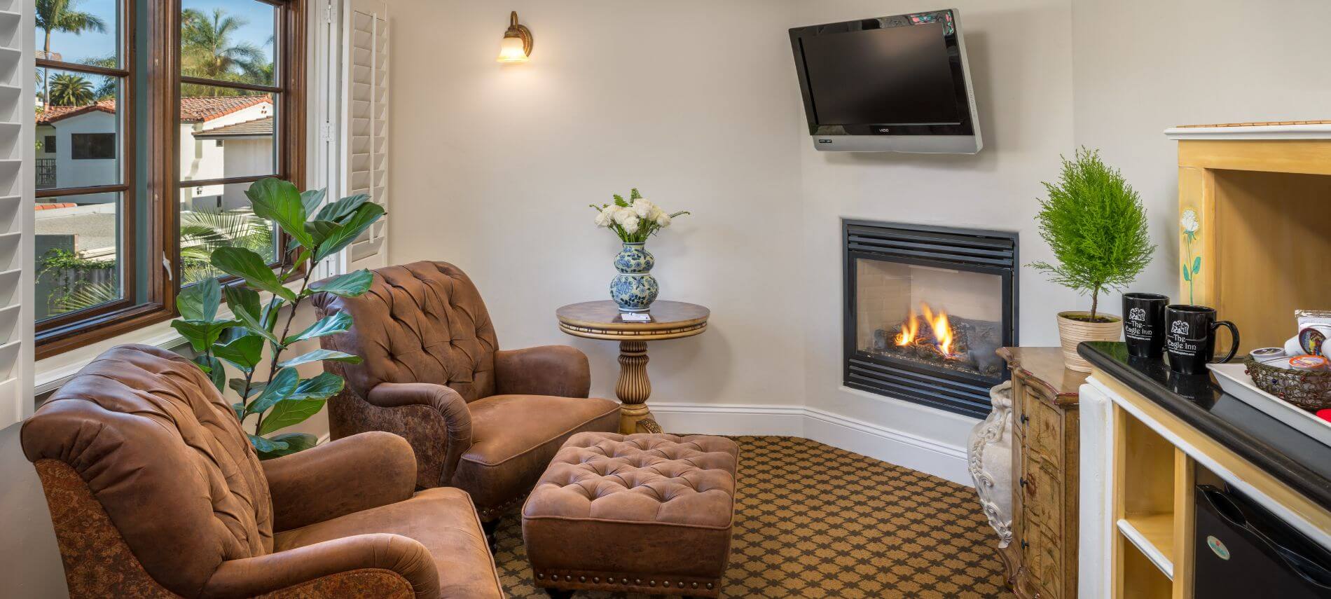 small room with flat tv hanging above fireplace, brown leather chairs with single ottoman, small shelves holding refreshments