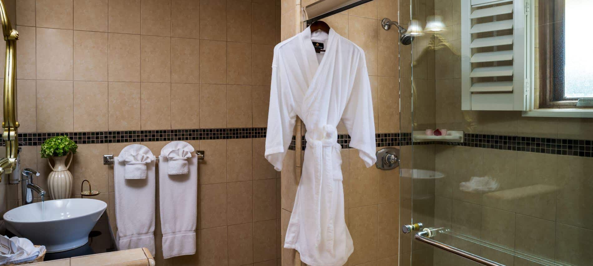 tan tiled bathroom with glass enclosed walk-in shower, white robe hanging, towels on rack, single sink