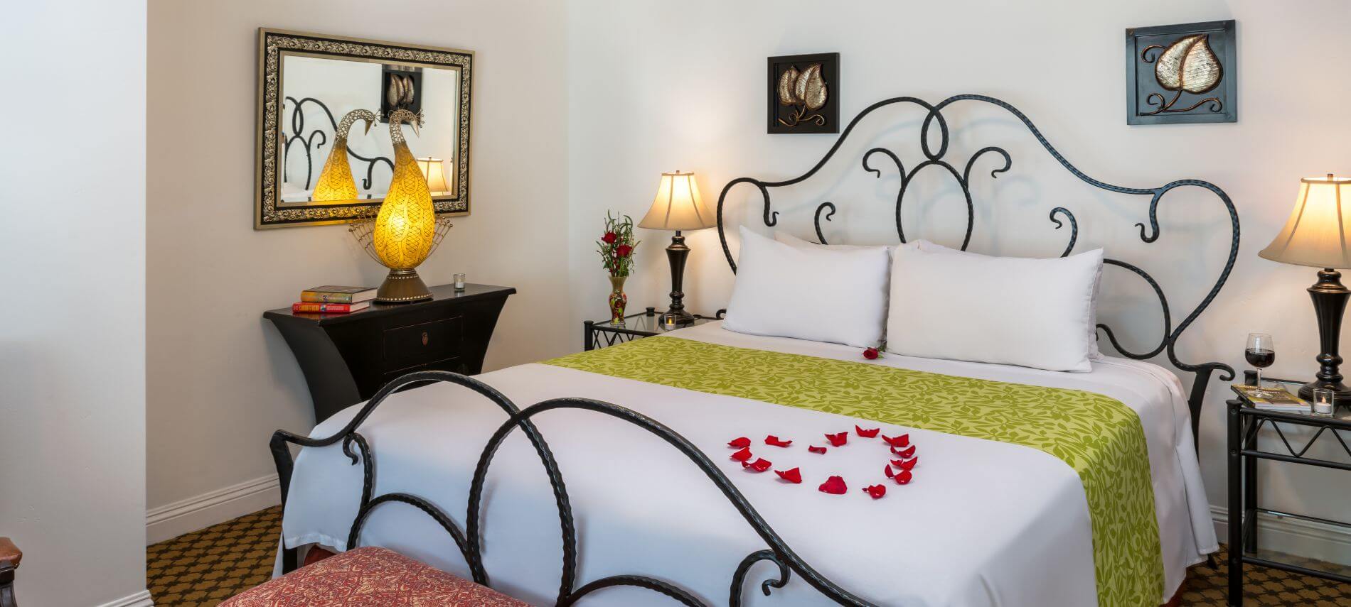 carpeted white room with wrought iron queen bed with white bedspread, stands with lamps, mirror and artwork hanging on walls