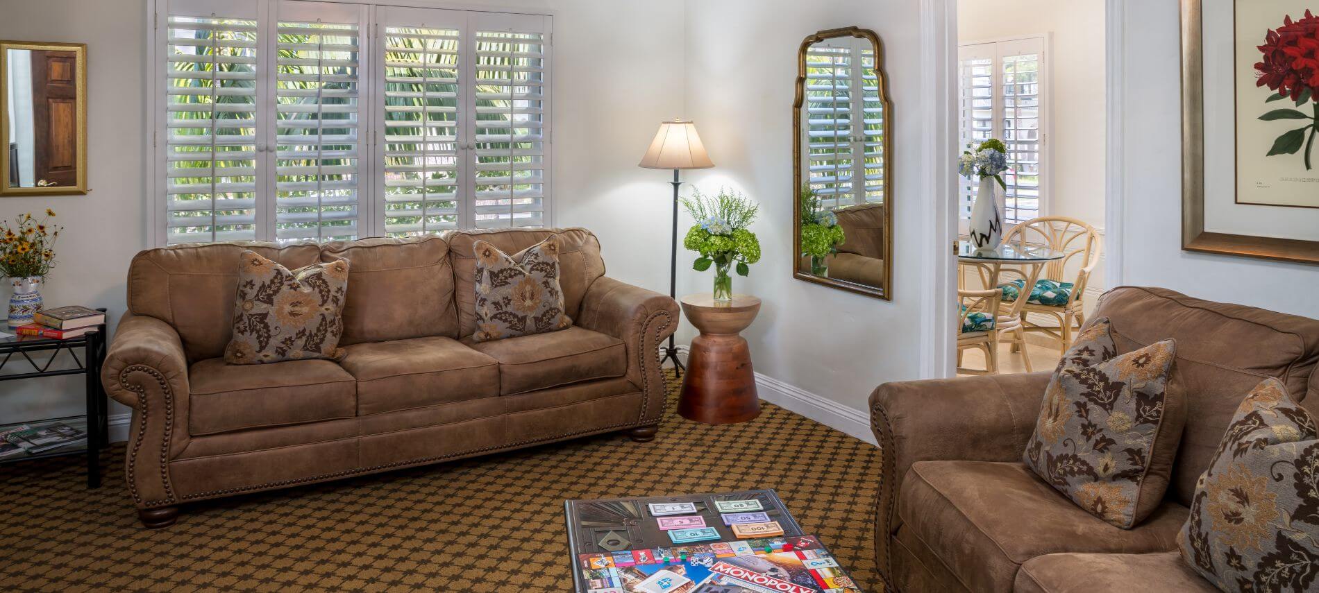 well lit carpeted living room with brown couches and throw pillows, shuttered white windows, view of kitchen through doorway