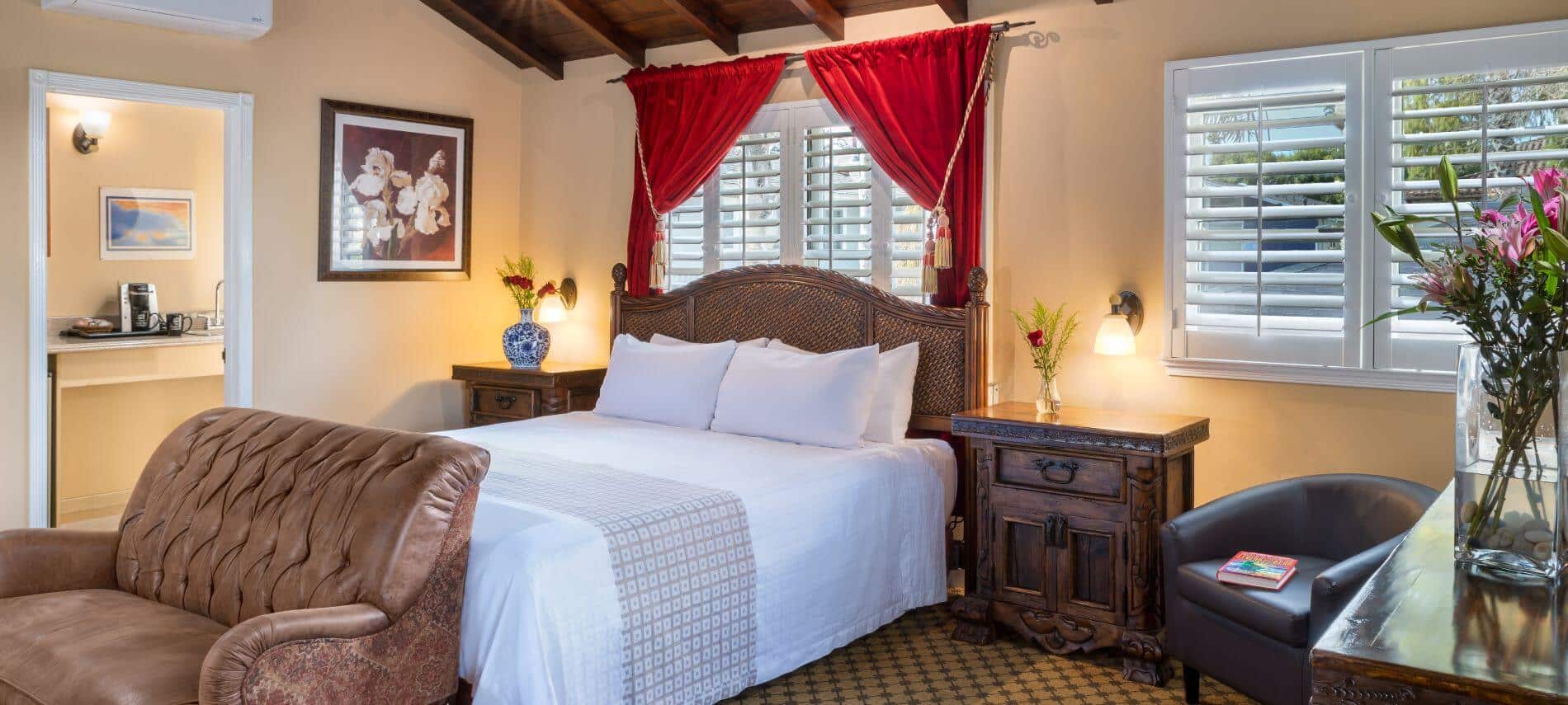 tan bedroom with queen bed with wicker headboard, red curtains behind bed, wood nightstands, brown leather loveseat, shuttered windows